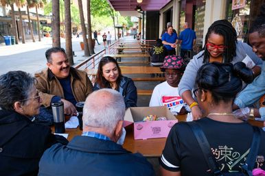 Smiling diverse group gathered around picnic tables on a sunny, palm-lined downtown street, sharing colorful sprinkled donuts from a pink box.