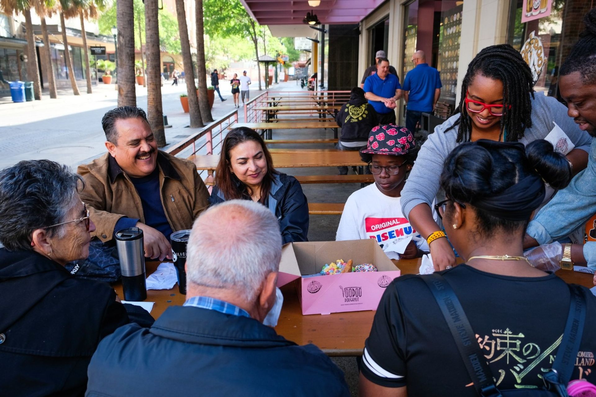 Smiling diverse group gathered around picnic tables on a sunny, palm-lined downtown street, sharing colorful sprinkled donuts from a pink box.