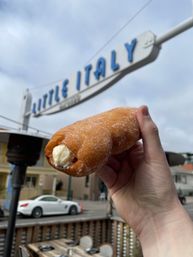 Close-up of a hand holding a sugar-dusted, cream-filled pastry roll against a cloudy sky with a blurred retro blue-and-white street sign, outdoor cafe seating and a white car in the background.