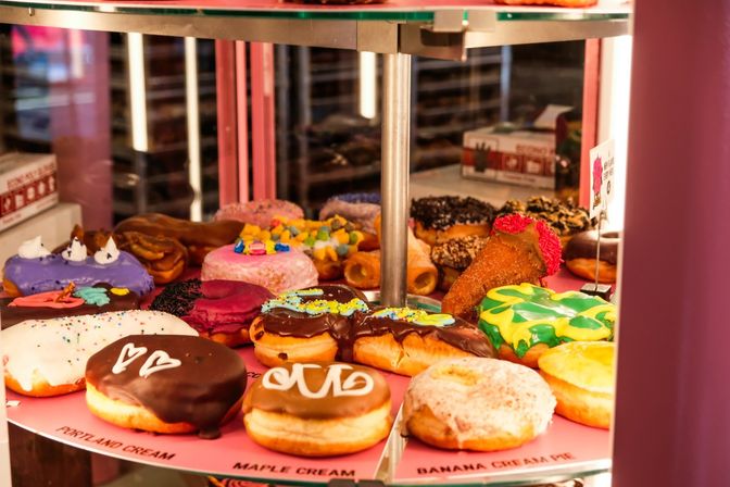 Colorful gourmet donuts with chocolate, icing, sprinkles and novelty toppings displayed on a pink rotating glass case in a bakery.