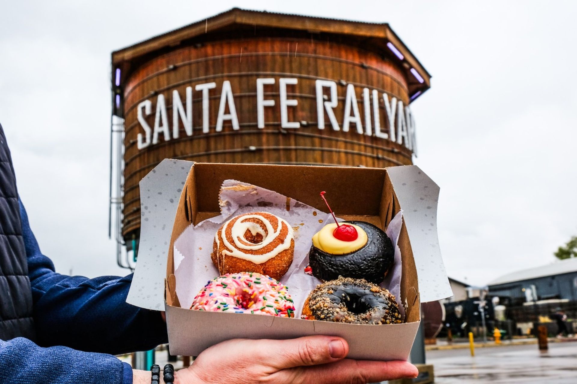 Box of four colorful assorted donuts held up in front of the Santa Fe Railyard wooden water tower on a cloudy day — pink sprinkle, cinnamon swirl, chocolate with cherry, and nut-topped