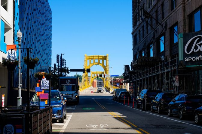 Downtown street with parked cars and storefronts framing a bright yellow suspension bridge over the river under a clear blue sky, with bike lane markings and hanging flower baskets.