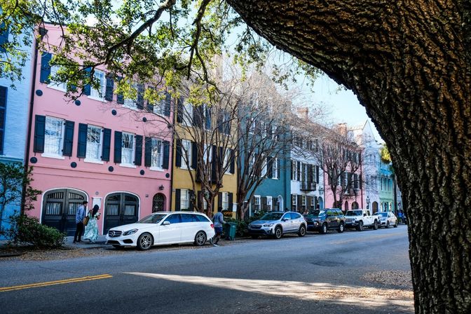 Oak tree-framed street view of colorful pastel row houses, bare winter trees, parked cars and pedestrians on a sunny historic downtown block