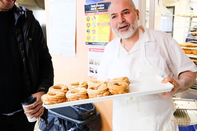 Smiling baker in a bakery kitchen holding a tray of fresh sugar‑coated crullers while a customer holds a takeaway coffee.
