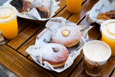Two powdered-sugar topped filled doughnuts with chocolate and vanilla centers on paper liners, served with a latte and two orange juices on a wooden outdoor table — breakfast pastry spread.