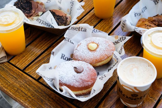Two powdered-sugar topped filled doughnuts with chocolate and vanilla centers on paper liners, served with a latte and two orange juices on a wooden outdoor table — breakfast pastry spread.