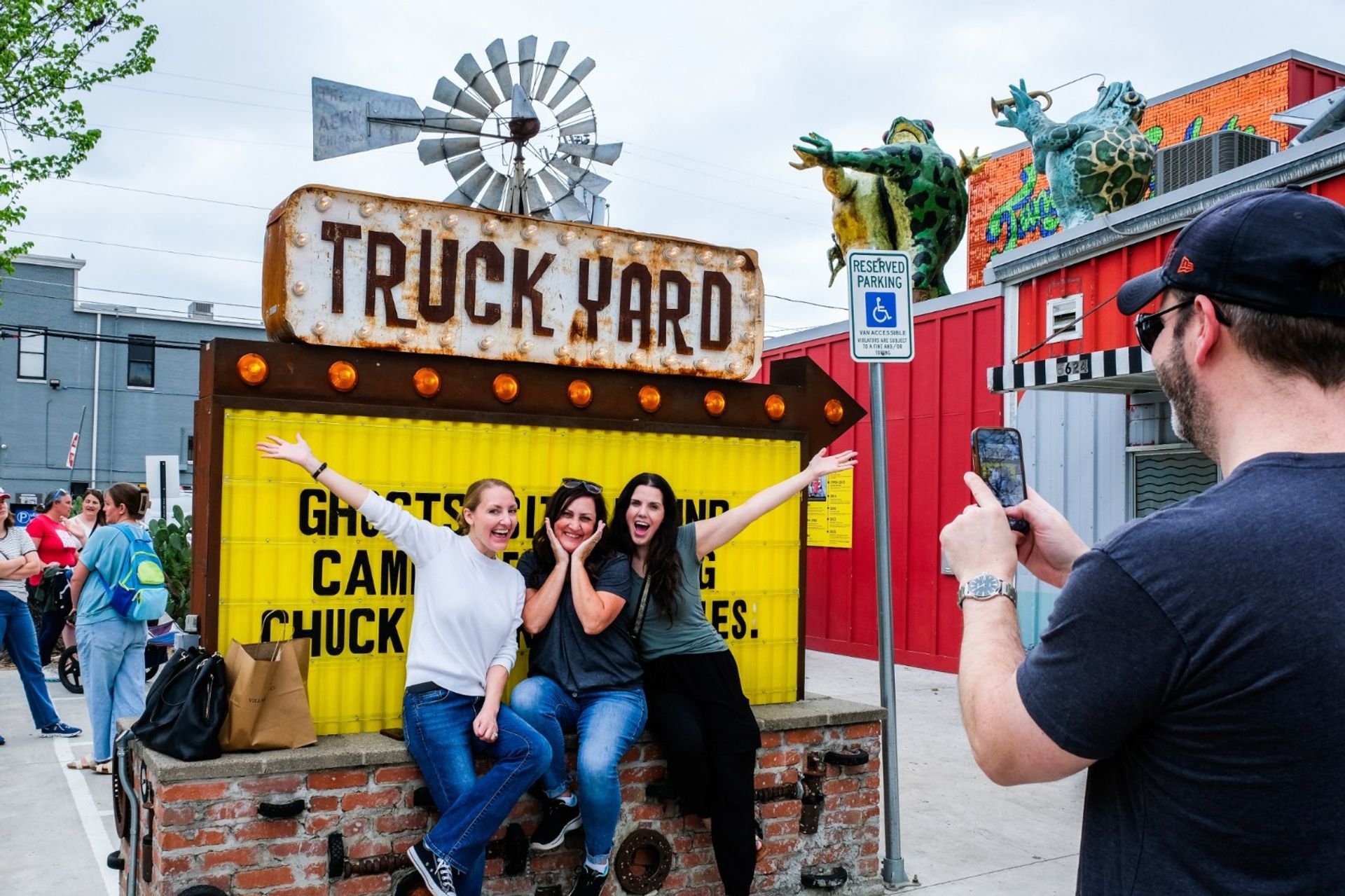 Three people cheer while sitting on a brick ledge beneath a weathered 'TRUCK YARD' marquee and yellow sign as a man photographs them with a smartphone in a colorful urban outdoor truck‑park with rooftop frog sculptures and a reserved parking sign.