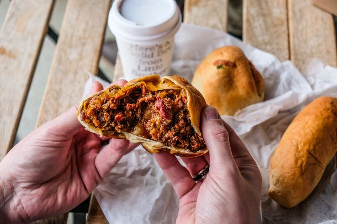 Close-up of a torn-open savory meat hand pie with shredded beef and melted cheese held over parchment on a rustic wooden table beside a takeaway coffee cup and extra baked pastries