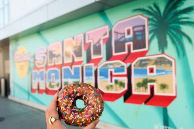 Hand holding a sprinkle-topped chocolate donut in front of a colorful Santa Monica mural depicting beach, pier and palm trees.