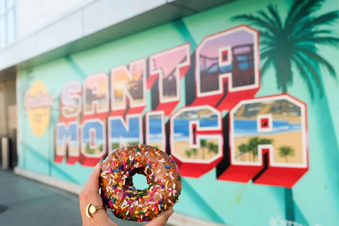 Hand holding a sprinkle-topped chocolate donut in front of a colorful Santa Monica mural depicting beach, pier and palm trees.