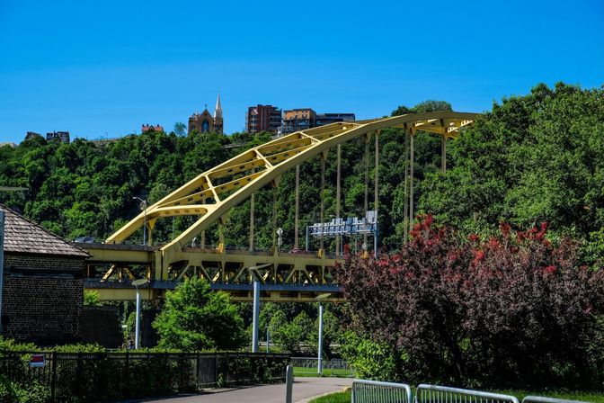 Bright yellow arched bridge crossing a leafy river valley with a church spire and apartment buildings on the hillside, paved path and colorful shrubs in the foreground under a clear blue sky.