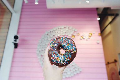 Close-up of a hand holding a chocolate-glazed donut topped with colorful rainbow sprinkles in front of a pink café wall with a blurred decorative swirl.