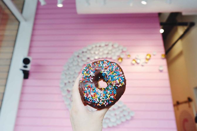 Close-up of a hand holding a chocolate-glazed donut topped with colorful rainbow sprinkles in front of a pink café wall with a blurred decorative swirl.