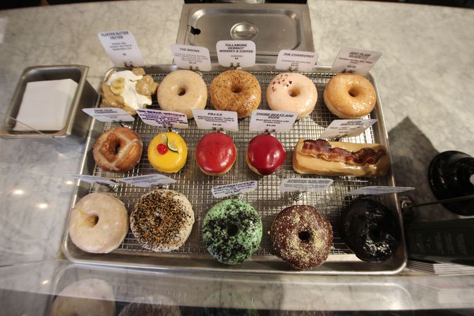 Assorted colorful artisanal donuts — glazed, frosted, sprinkled and a bacon-topped bar — arranged on a wire rack inside a glass bakery display on a marble countertop.