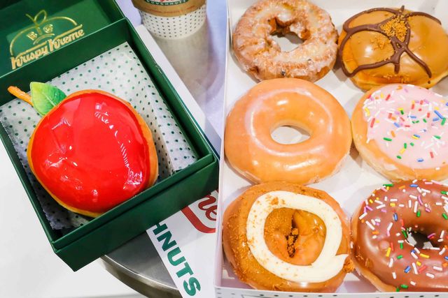 Shiny red apple-shaped donut in a green pastry box beside a white box of assorted glazed, chocolate, and sprinkle-topped donuts with a coffee cup on a café table
