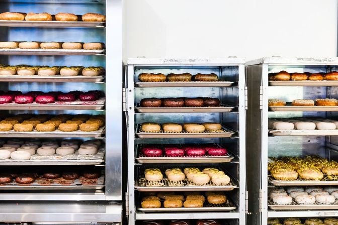 Assorted glazed and topped donuts on metal trays in a commercial bakery kitchen - rows of chocolate, pink berry, powdered and nut-topped varieties cooling on baking racks.