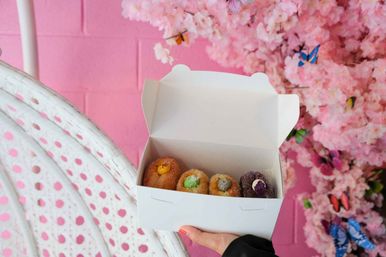 Hand holding a white takeout box with four colorful filled mini doughnuts against a pink wall and cherry-blossom display with decorative butterflies.