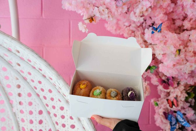Hand holding a white takeout box with four colorful filled mini doughnuts against a pink wall and cherry-blossom display with decorative butterflies.
