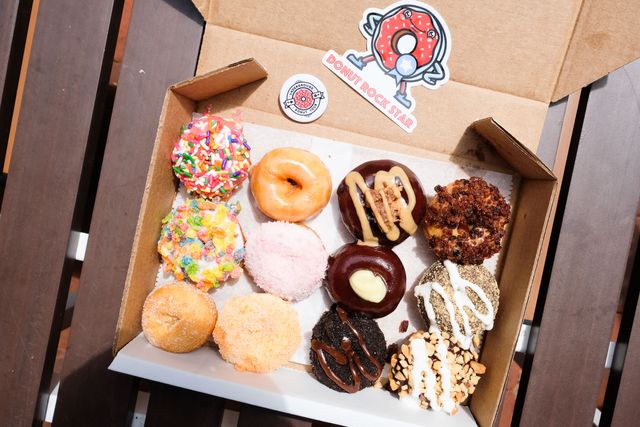 Assorted mini gourmet donuts — colorful sprinkles, glazed, chocolate ganache, nut and cereal toppings — in a cardboard takeout box on a wooden outdoor bench