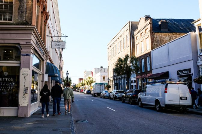 Sunlit historic downtown street in a southern coastal town with brick storefronts and awnings, palm trees lining the sidewalk, parked cars and a white van, and three pedestrians walking
