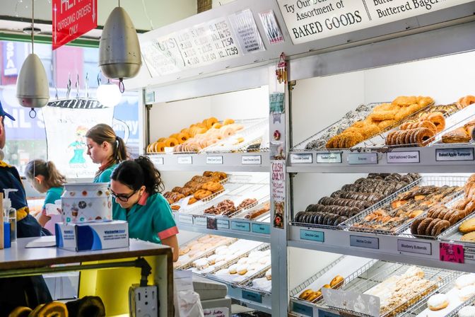 Busy donut shop display case filled with assorted fresh donuts and pastries, staff serving customers behind the counter