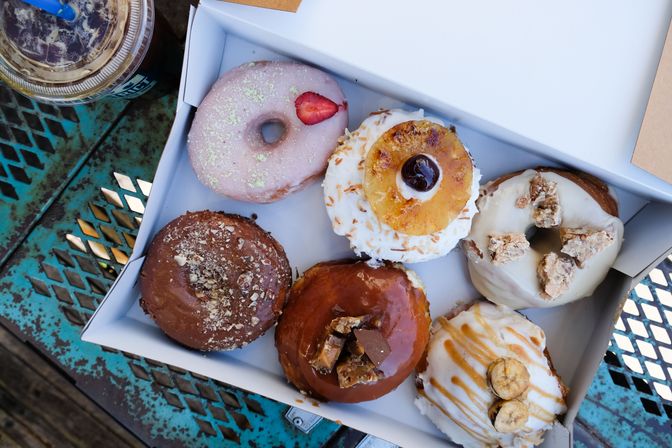 Box of six assorted gourmet donuts with strawberry, grilled pineapple and cherry, white crumble, chocolate and caramel toppings, and banana-caramel, on a teal outdoor café metal table next to an iced coffee