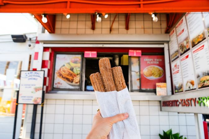 Crispy cinnamon-sugar churros wrapped in paper, held up in front of a bright outdoor street-food window and menu boards