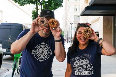 Two adults on a downtown sidewalk playfully holding sprinkled, glazed, and drizzled donuts over their eyes, smiling and wearing donut-themed t-shirts during a daytime urban donut tour.