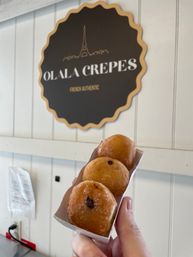 Hand holding a small tray of three sugared jam-filled mini doughnuts in front of a French crepe shop sign with an Eiffel Tower logo on a white shiplap wall.