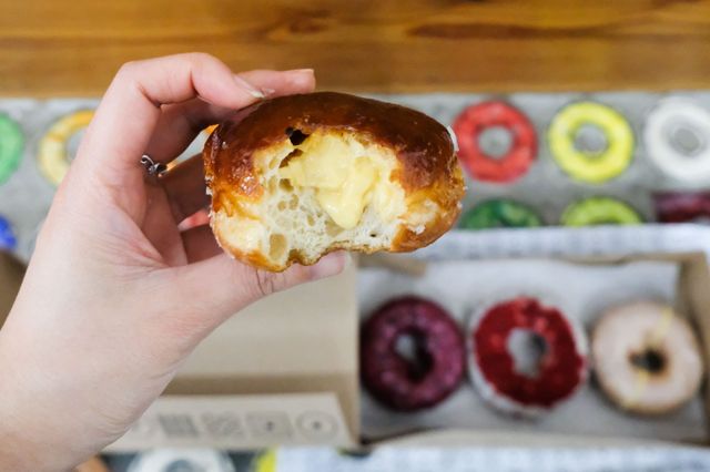 Hand holding a bitten glazed custard-filled donut with creamy yellow filling over a box of colorful frosted donuts on a bakery countertop