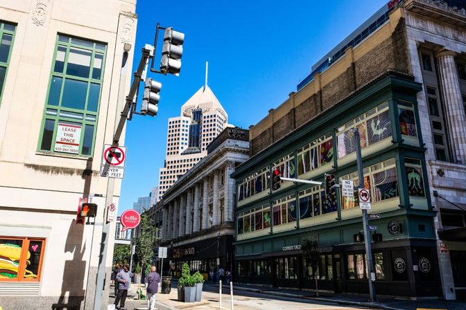 Vibrant downtown street scene with a columned neoclassical building and green corner storefront adorned with colorful window art, traffic lights and street signs, pedestrians on the sidewalk, and a pointed modern skyscraper against a bright blue sky.