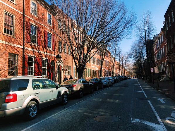Sunlit tree-lined urban street with historic red brick rowhouses, parked cars along the curb, bare winter trees casting branch shadows under a clear blue sky.
