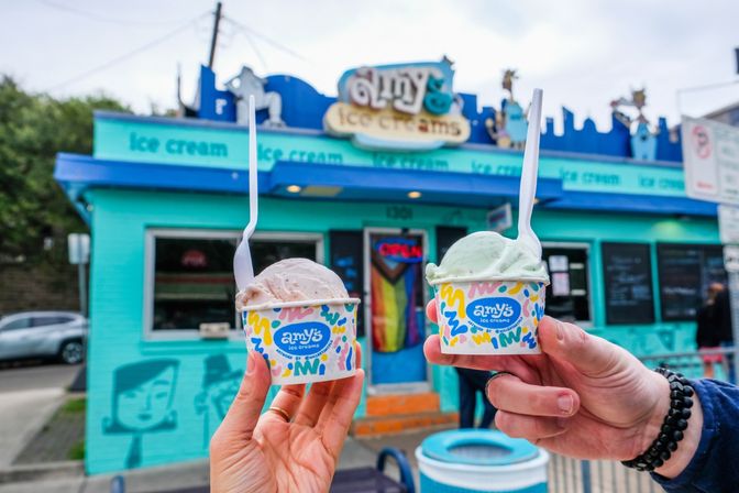 Two hands holding colorful paper cups of scooped ice cream with plastic spoons in front of a bright turquoise ice cream shop featuring a rainbow-striped doorway and neon OPEN sign.