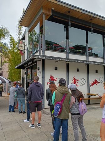 People queued outside a modern two-story donut and coffee shop with a glass upper patio, white exterior featuring red devil-and-donut murals, and outdoor seating on a busy urban sidewalk.