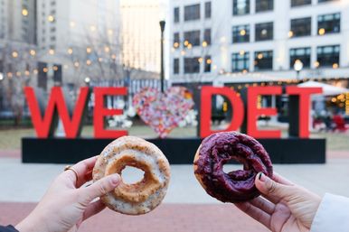 Two hands holding a poppy-seed glazed donut and a berry-glazed donut in front of a large red "WE ♥ DET" public art sign in a downtown Detroit plaza with string lights and office buildings in the background