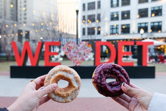 Two hands holding a poppy-seed glazed donut and a berry-glazed donut in front of a large red "WE ♥ DET" public art sign in a downtown Detroit plaza with string lights and office buildings in the background
