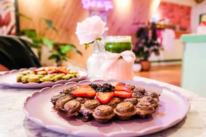 Close-up of a strawberry-topped bubble waffle on a pink scalloped plate with a green matcha drink, small vase with a pink rose, and a cozy cafe background