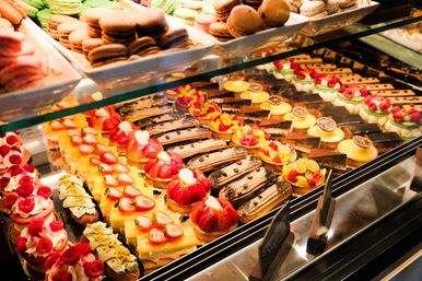 Patisserie-style glass display case packed with colorful pastries — green and brown macarons, strawberry and raspberry fruit tarts, chocolate éclairs and lemon custard tarts.