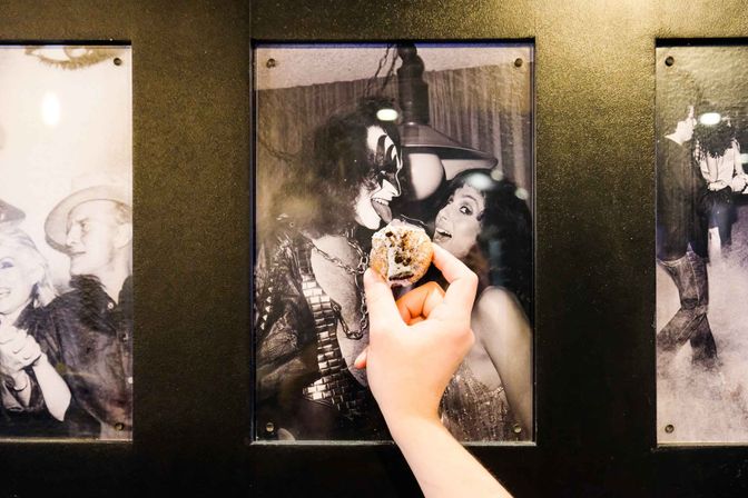 Hand holding a sugared pastry up to a framed black-and-white photo of a costumed rock performer and a laughing woman on a black gallery wall.