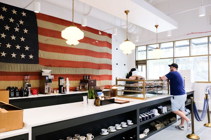 Bright neighborhood coffee shop and bakery interior with pastry display case, oversized vintage American flag on the wall, hanging pendant lights, and a casually dressed customer at the counter.