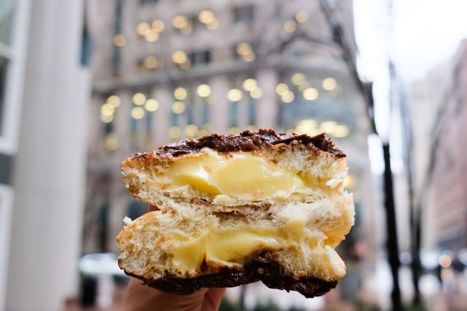 Creamy custard oozing from a chocolate-topped pastry bun held against a blurred city street with warm bokeh lights.