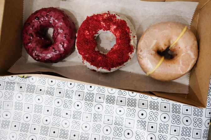 Top-down view of three colorful donuts in a cardboard box on a patterned table: berry-glazed donut, white-glazed donut with red crumb topping, and a classic glazed donut with yellow drizzle.