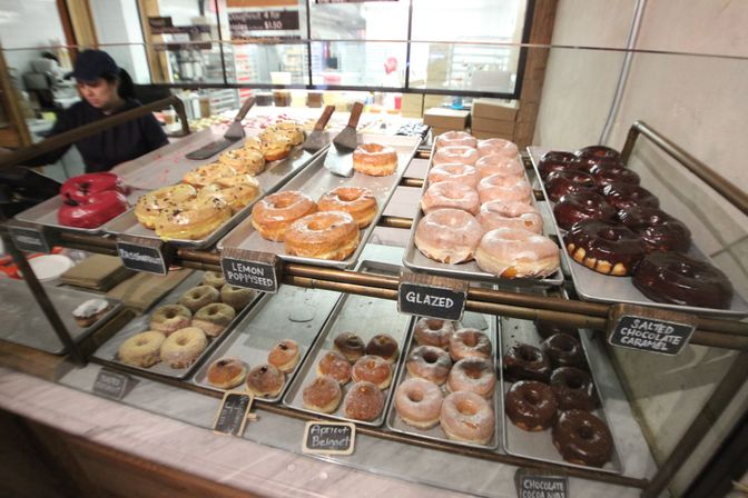Assorted donuts in a glass bakery display case — rows of glazed, chocolate, lemon poppyseed and apricot-filled beignet-style pastries with small chalkboard labels on trays.