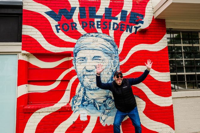 Man wearing sunglasses and cap poses with arms raised in front of a bright red-and-white swirled brick street mural featuring a blue portrait in a bandana and the bold text "WILLIE FOR PRESIDENT"
