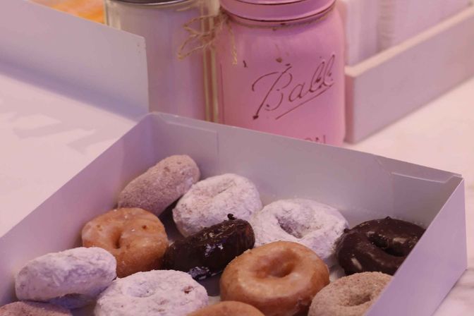 Assorted mini donuts (powdered, glazed, chocolate) in a white bakery box on a table, with a pink painted mason jar in the background.