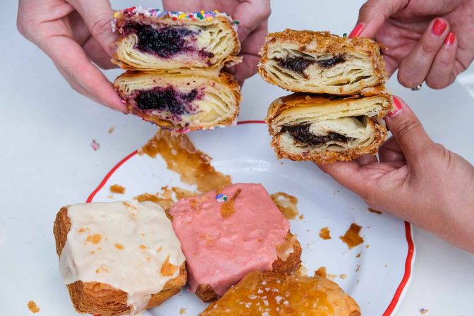 Hands hold halved flaky croissant-style pastries revealing blueberry jam and chocolate fillings on a white plate with red rim, surrounded by square glazed pastries with pink and white frosting and sprinkles — bakery-style brunch treat.