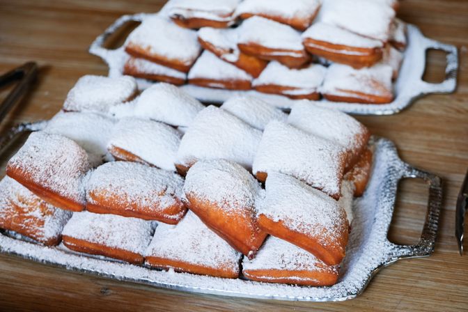 Puffy golden beignets dusted with powdered sugar, neatly stacked on metal serving trays on a wooden table