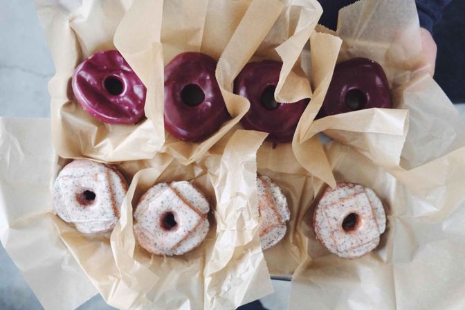 Top-view of six assorted donuts in a takeout box: three glossy dark purple glazed donuts and three speckled vanilla glazed donuts, each nestled in parchment paper.