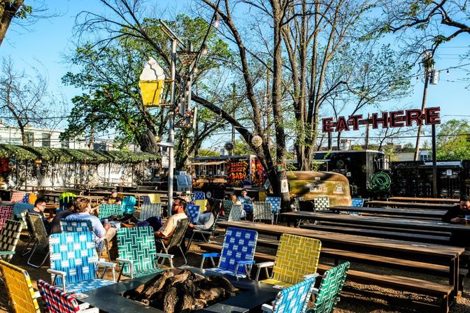 Sunny outdoor urban beer-garden and food-truck patio with colorful woven lawn chairs around a fire pit, long wooden picnic tables, trees overhead, neon "EAT HERE" sign and a vintage ice-cream cone sign.