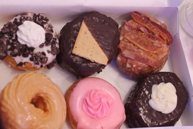 Assorted gourmet donuts in a bakery box: cookies-and-cream with whipped topping, chocolate-covered with graham triangle, maple bacon, glazed cruller, pink frosted with a swirl, and chocolate topped with cream.